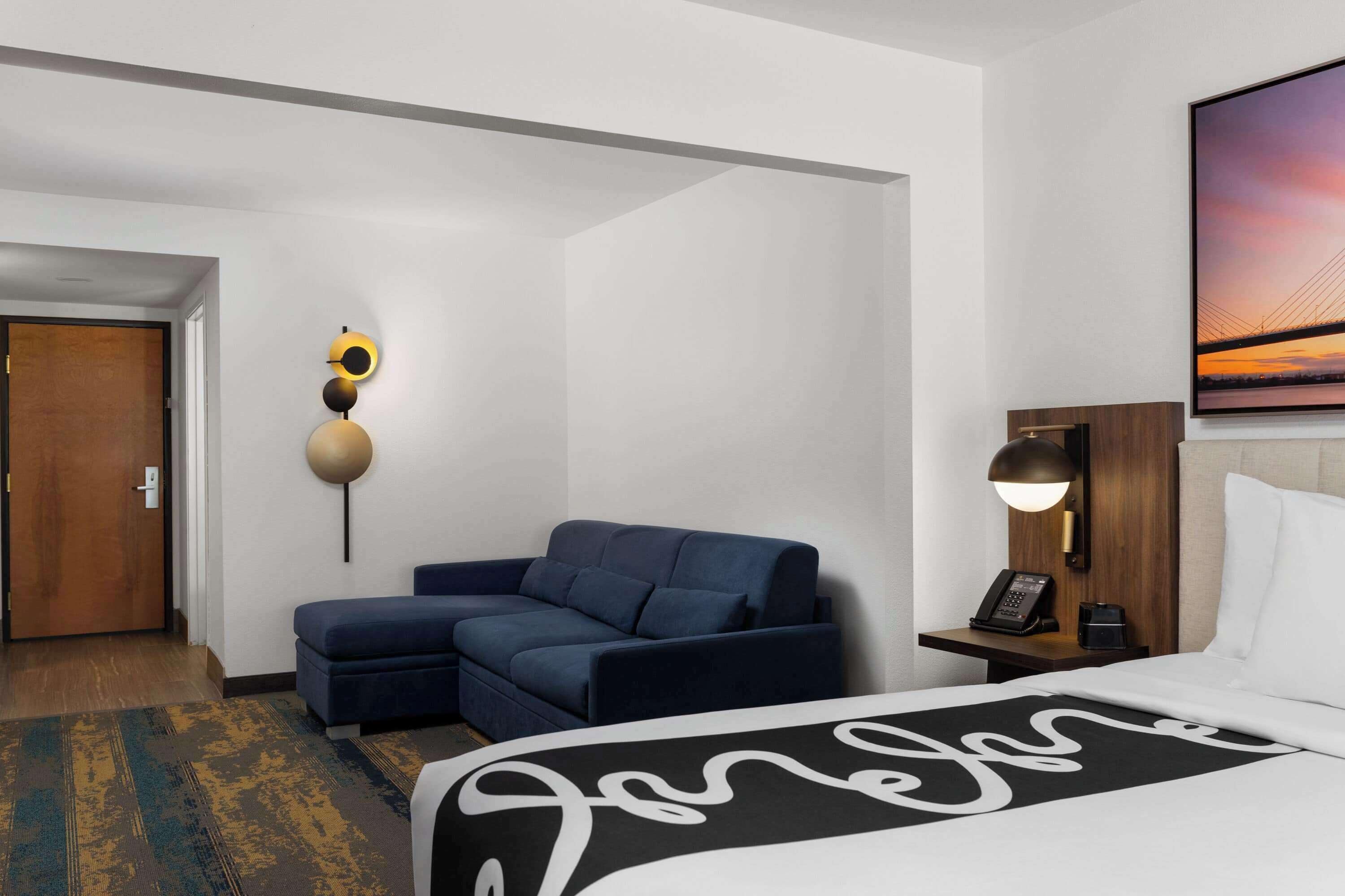 A hotel room with a bed, a sofa, a nightstand with a lamp, a wall-mounted phone, and a painting of a sunset.