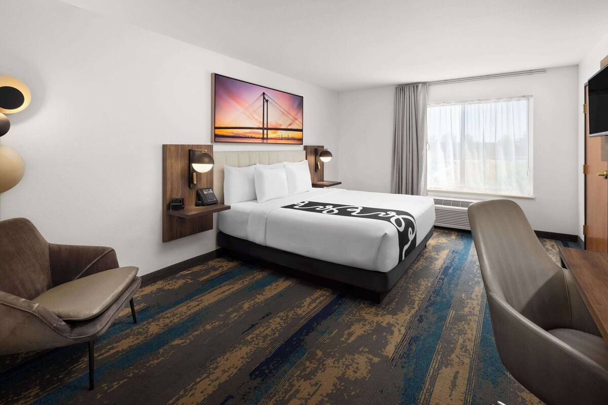 A hotel room with a bed, two chairs, a desk, a painting of a bridge, and a large window with curtains.