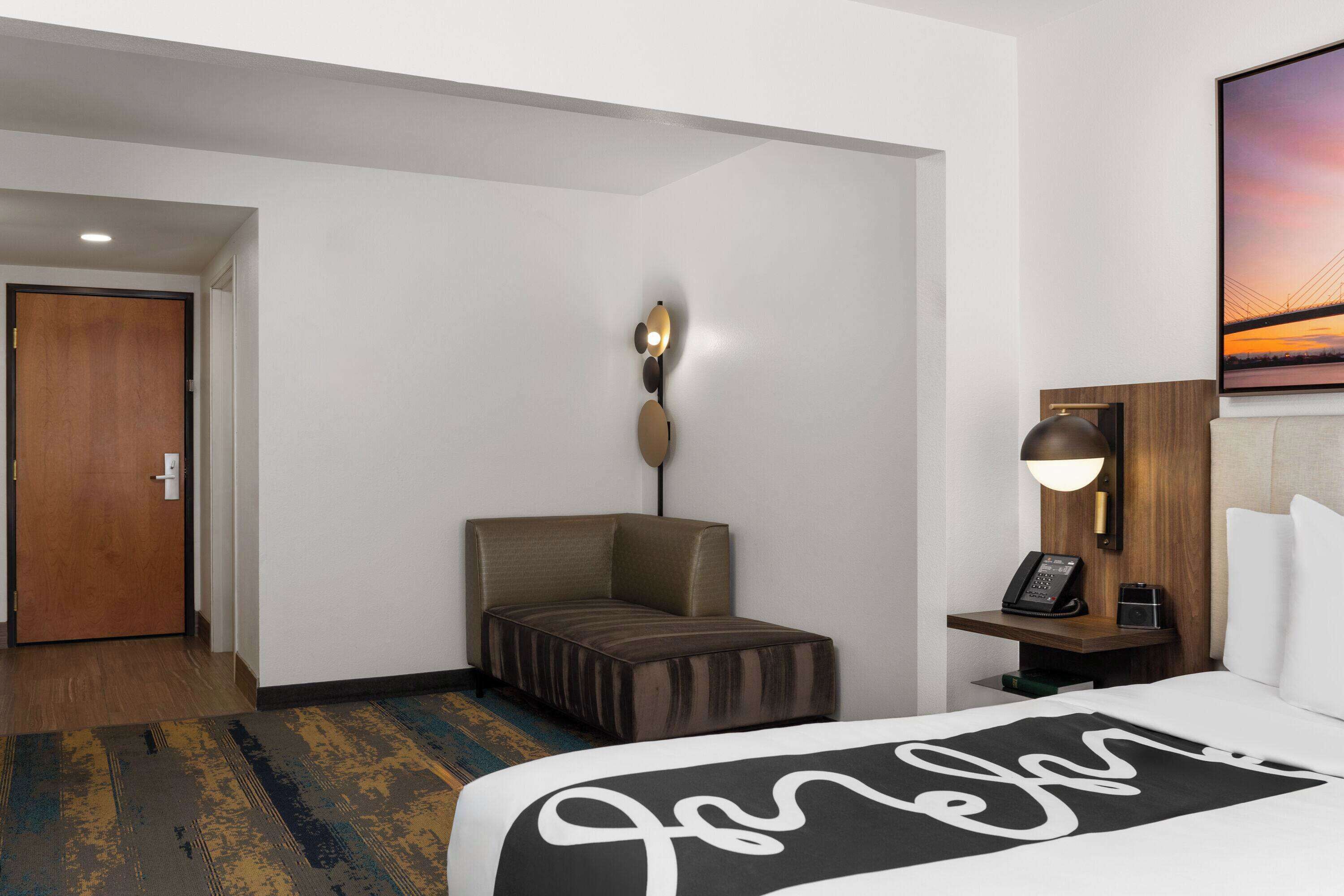 A hotel room with a bed, a nightstand with a lamp, a seating area, and a door.