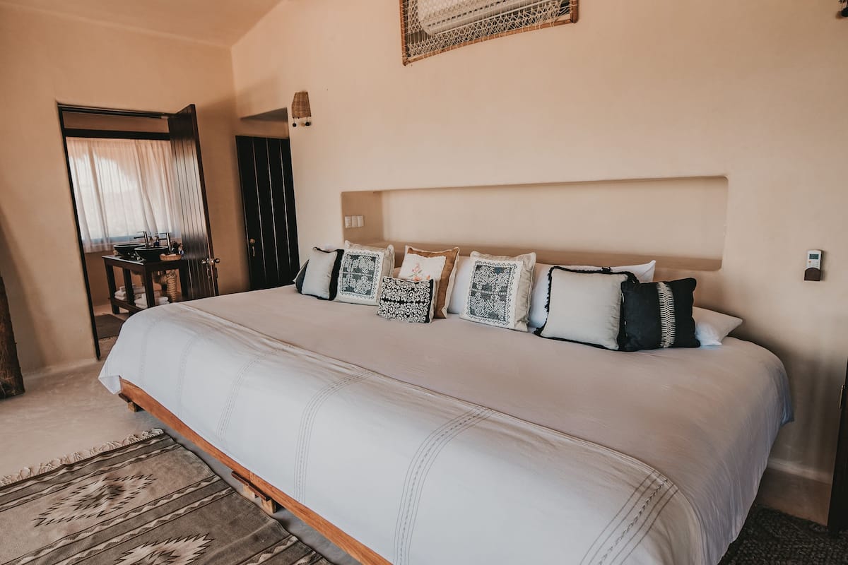 Uma cama grande com colcha branca e várias almofadas, cabeceira de madeira, tapete tecido e ventilador de teto.