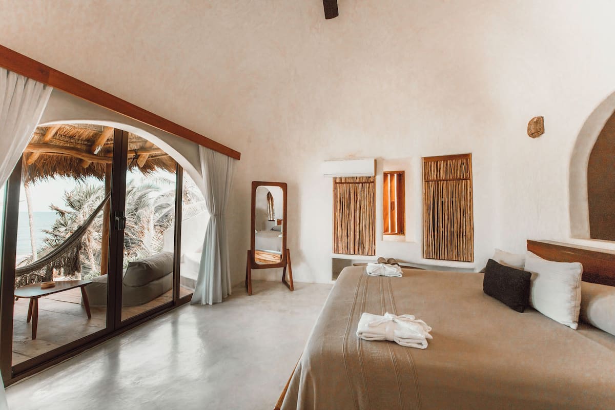 Quarto com cama grande, cabeceira de madeira, espelho e vista para palmeiras através de uma porta de vidro.