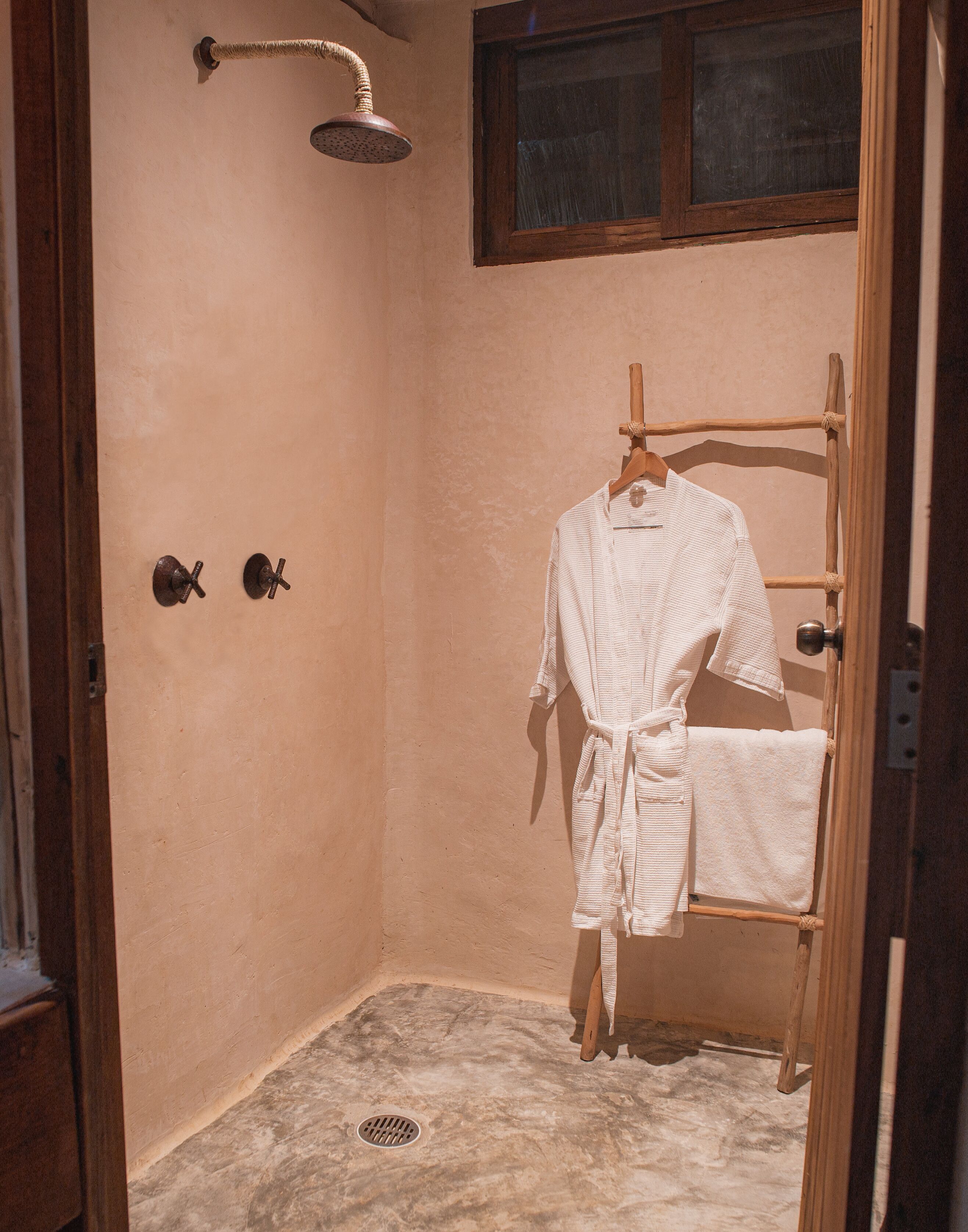A small, enclosed shower area with a wooden rack holding a bathrobe and towel, a showerhead, and a window above.