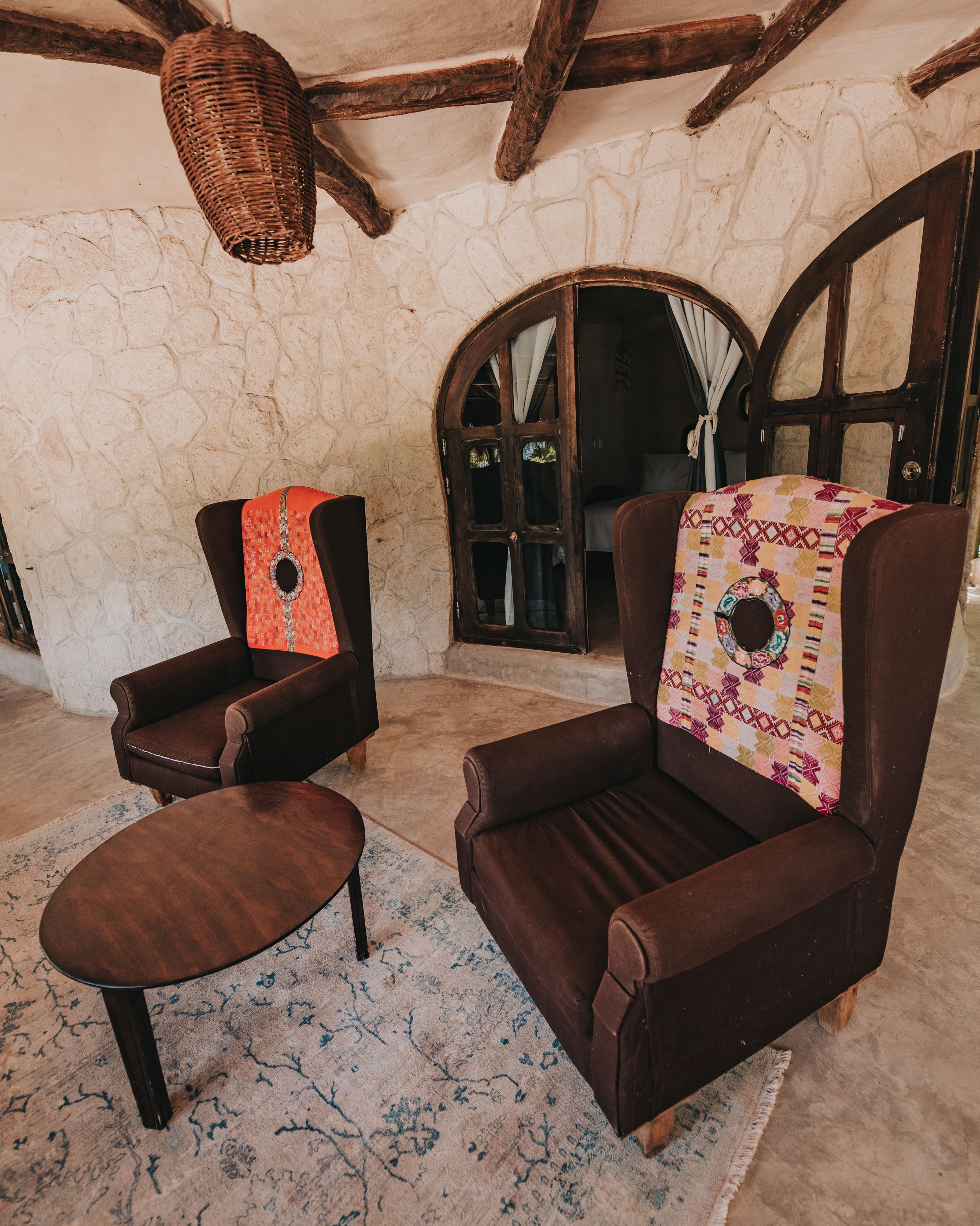 A cozy room with two brown leather armchairs, a small wooden table, and a patterned throw on one chair. A stone wall and wooden beams are visible in the background.