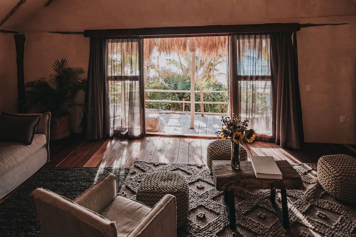 Um quarto aconchegante com teto inclinado, uma janela grande com cortinas, um sofá, uma mesa com um vaso e uma planta em vaso.