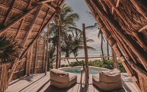 Una terraza de madera con dos puffs color beige, una piscina pequeña y vista al océano y palmeras.