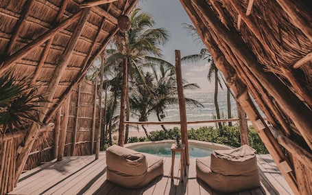 A wooden deck with two beige bean bags, a small pool, and a view of the ocean and palm trees.