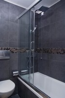 Ένα σύγχρονο μπάνιο με ένα ντους που διαχωρίζεται με γυάλινο διάφραγμα, μια λευκή τουαλέτα και σκούρα πλακάκια στους τοίχους.