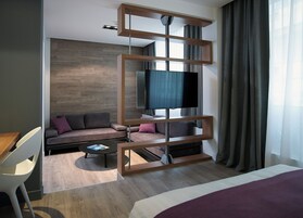 Quarto de hotel moderno com chão de madeira, um sofá com almofadas, uma mesa de centro, uma TV fixada em uma estrutura de madeira e uma cama com manta roxa.