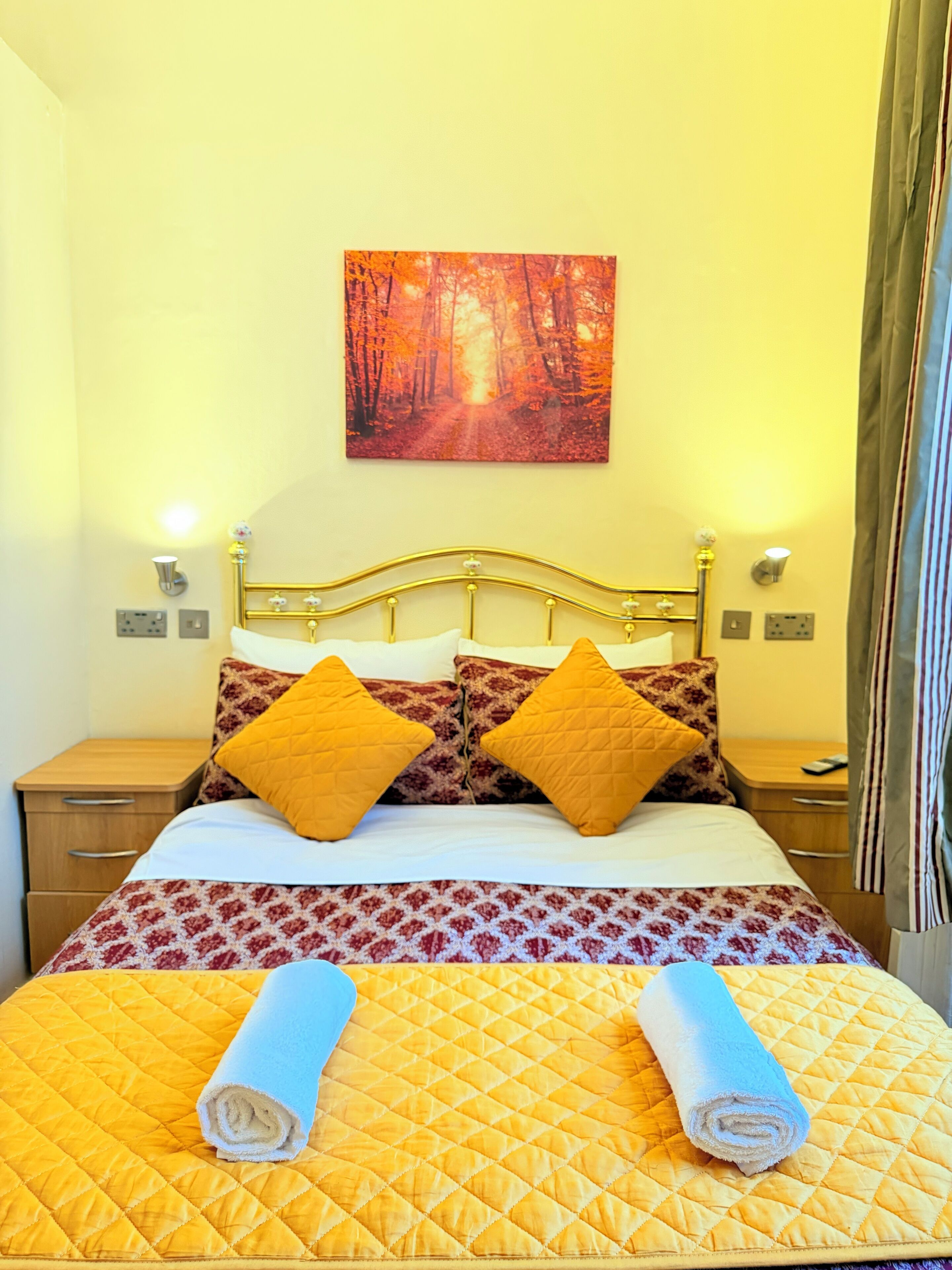 A neatly made bed with a patterned bedspread and two pillows, a painting of a forest on the wall, and a nightstand with a lamp.