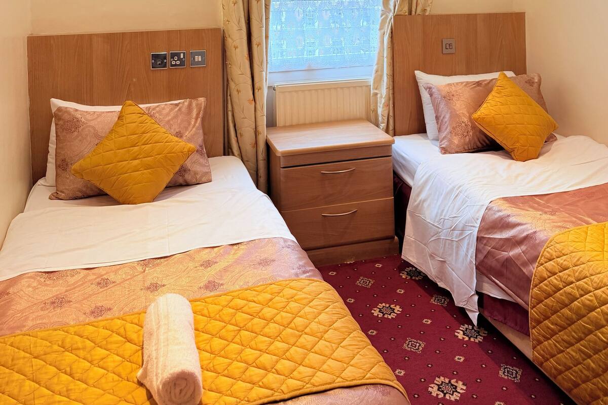 A hotel room with two beds, a wooden headboard, a small wooden nightstand, a window with curtains, and a radiator.