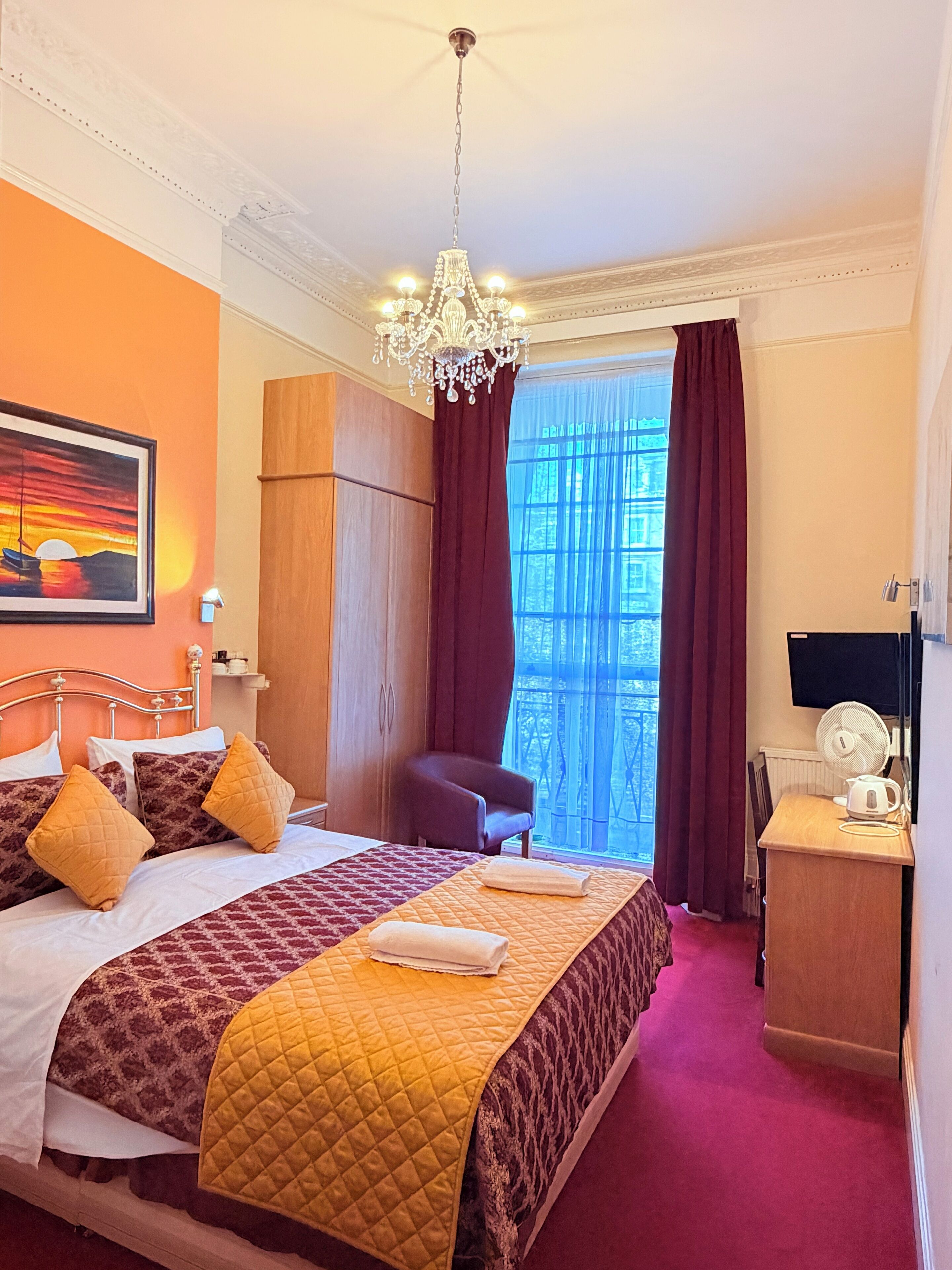 A hotel room with a bed, a desk, a chair, a fan, and a painting on the wall.