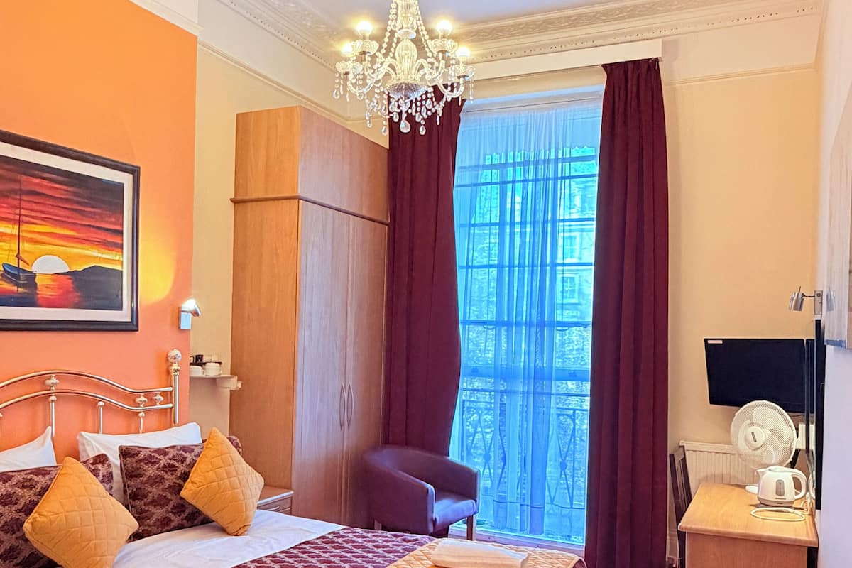 A hotel room with a bed, a desk, a chair, a fan, and a painting on the wall.