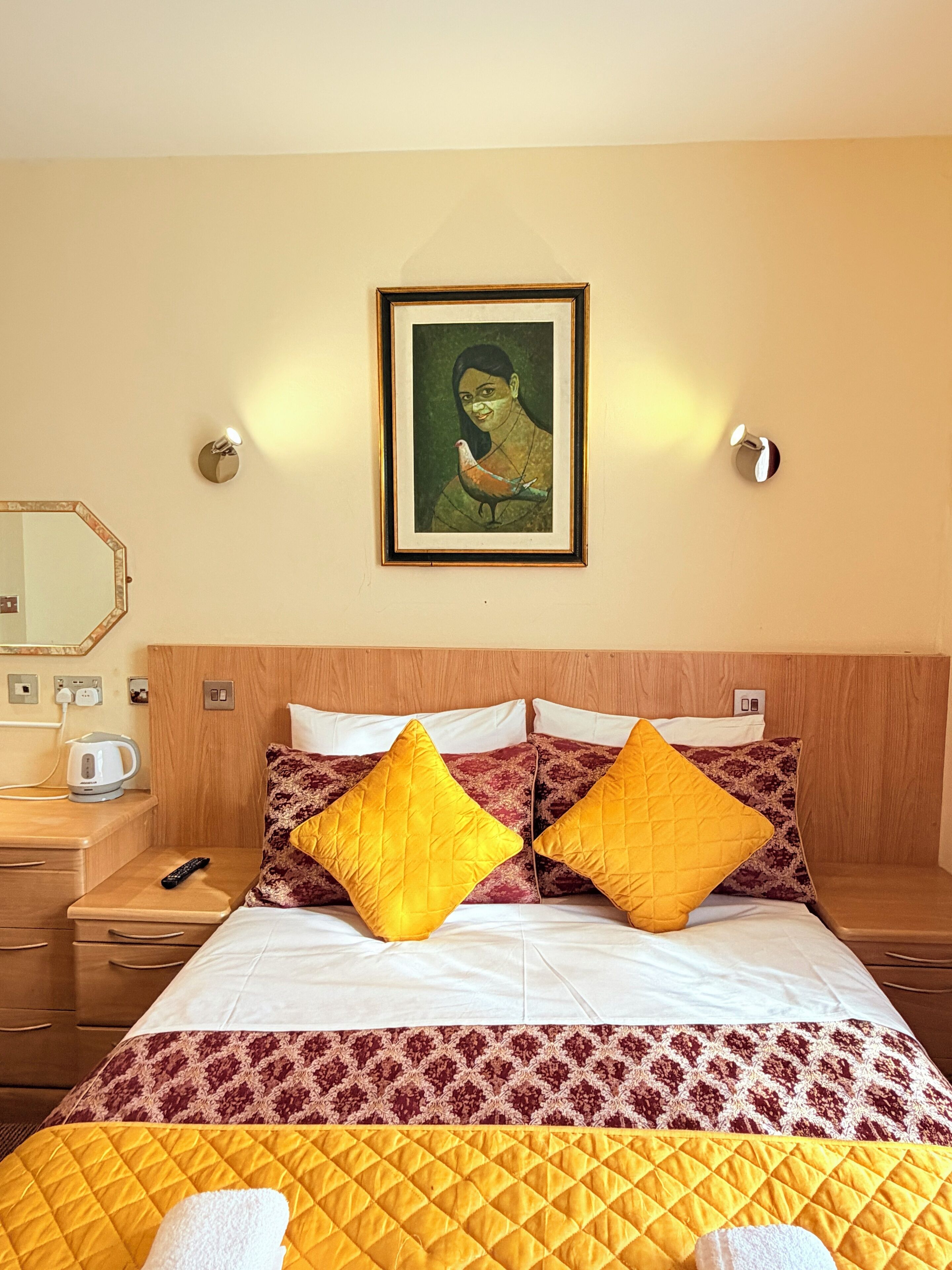 A hotel room with a bed, two pillows, a painting on the wall, a nightstand, a kettle, and a remote control.