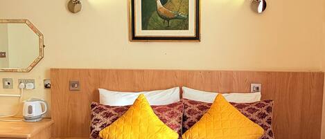 A hotel room with a bed, two pillows, a painting on the wall, a nightstand, a kettle, and a remote control.