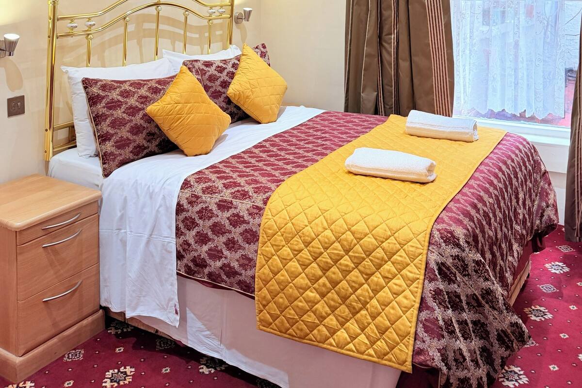A neatly arranged hotel room with a bed, a bedside table, a nightstand, a window with curtains, and a painting on the wall.