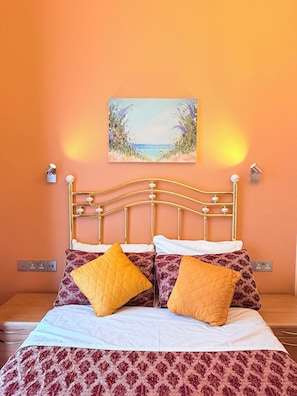 A bed with a patterned bedspread, two pillows, a gold headboard, and a painting of a beach scene on the wall. - Albro House Hotel (London)