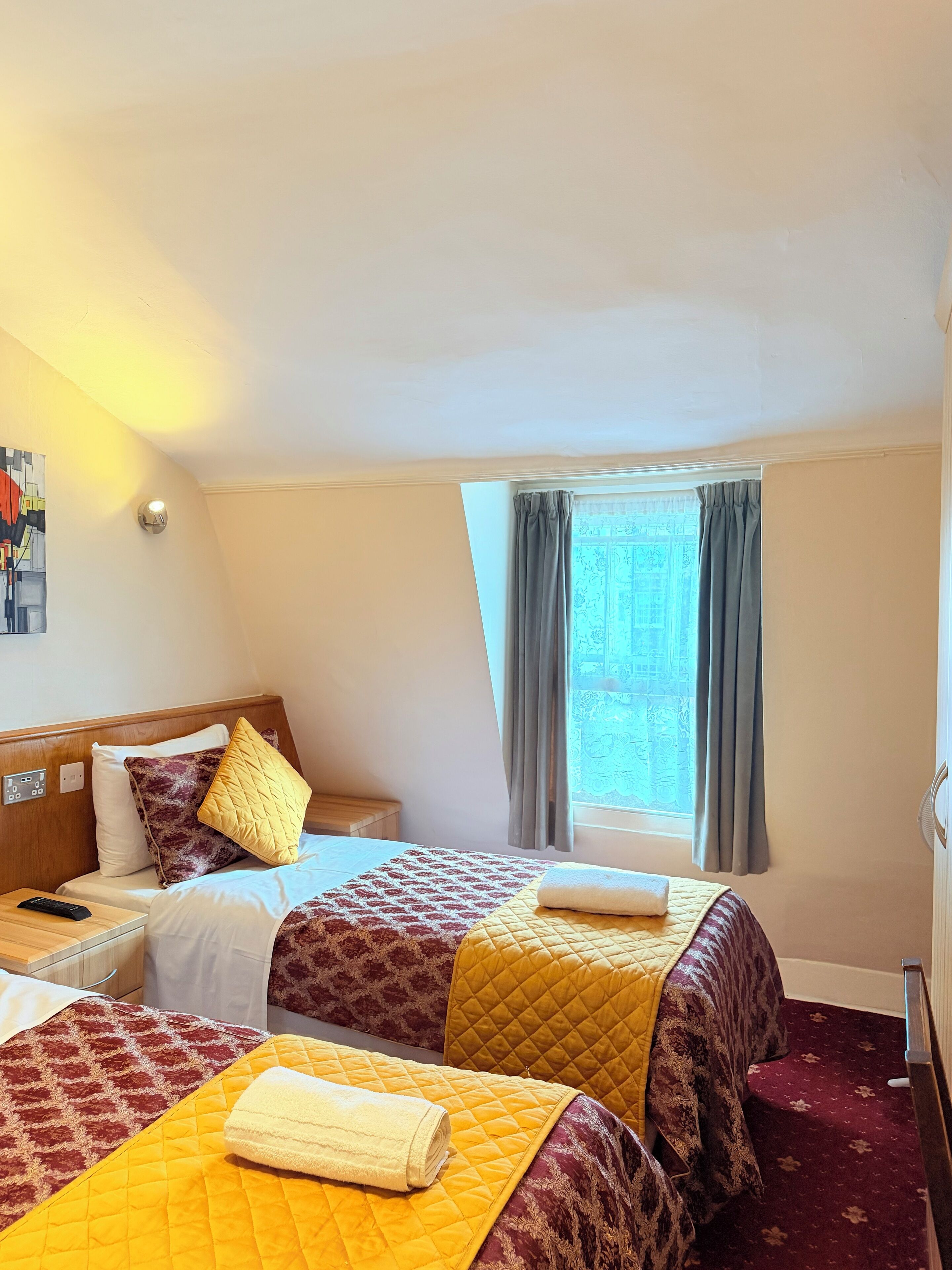 A hotel room with two beds, a nightstand, a painting on the wall, and a window with curtains.