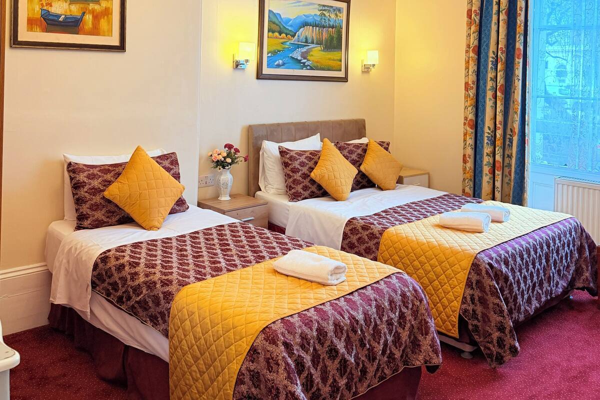 A hotel room with two beds, a chandelier, framed paintings, and a window with curtains.