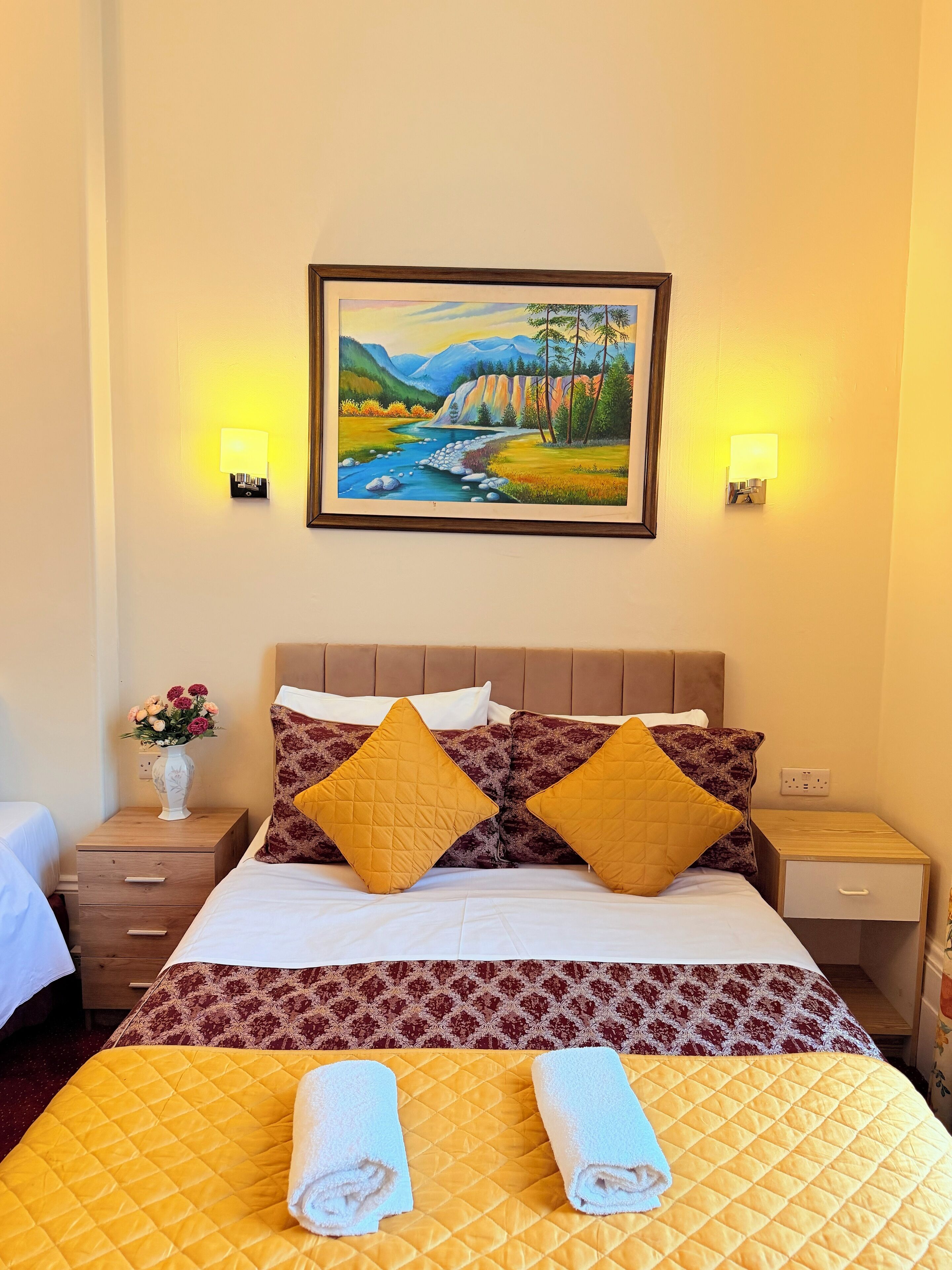 A hotel room with a bed, two nightstands, a painting, and a vase of flowers.