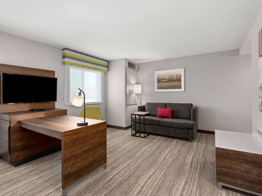 Una habitación de hotel moderna con un escritorio de madera, un televisor de pantalla plana, un sofá gris y una pequeña mesita.
