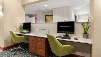Zona de recepción de hotel con dos mostradores, cada uno con computadora, teclado y ratón. Hay dos sillas verdes, un mueble de madera con cajones y dos plantas en macetas.