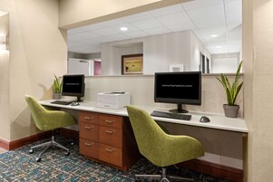 Zona de recepción de hotel con dos mostradores, cada uno con computadora, teclado y ratón. Hay dos sillas verdes, un mueble de madera con cajones y dos plantas en macetas.
