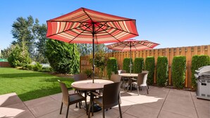 A patio with round tables and chairs, red striped umbrellas, and a wooden fence.
