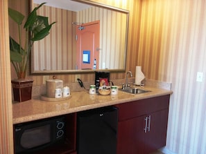 Una cocina con microondas, cafetera y lavabo. Hay un espejo sobre el lavabo y una planta en maceta en la encimera.