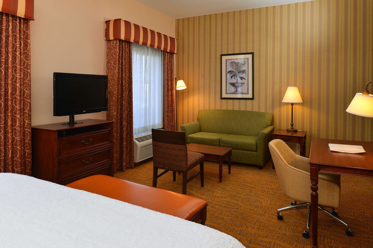 A hotel room with a bed, a desk, a green armchair, a wooden dresser, a television, and a window with curtains.