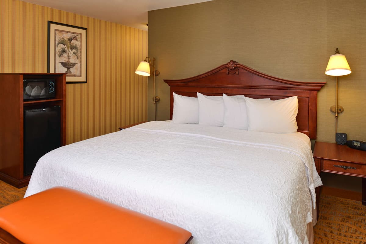 A hotel room with a large bed, a wooden headboard, two bedside lamps, a small desk with a microwave, and a framed picture on the wall.