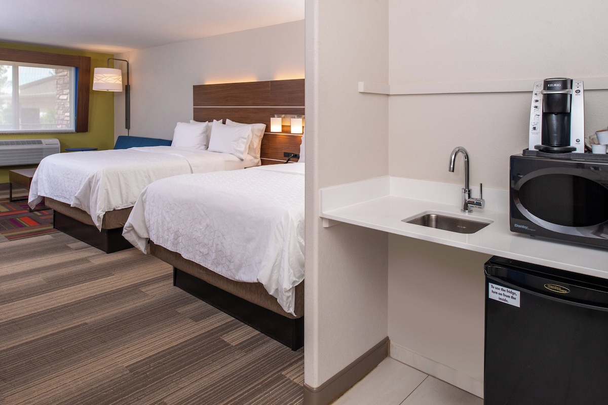 A hotel room with two beds, a desk, a window, a kitchenette with a coffee maker, and a trash bin.