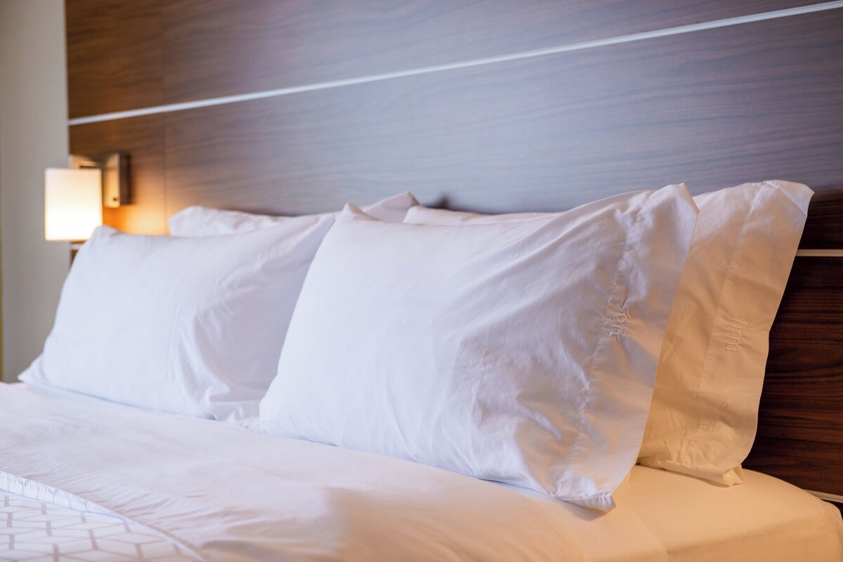 A neatly made bed with white linens and pillows, a wooden headboard, and a wall-mounted lamp.