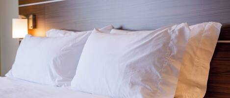A neatly made bed with white linens and pillows, a wooden headboard, and a wall-mounted lamp.