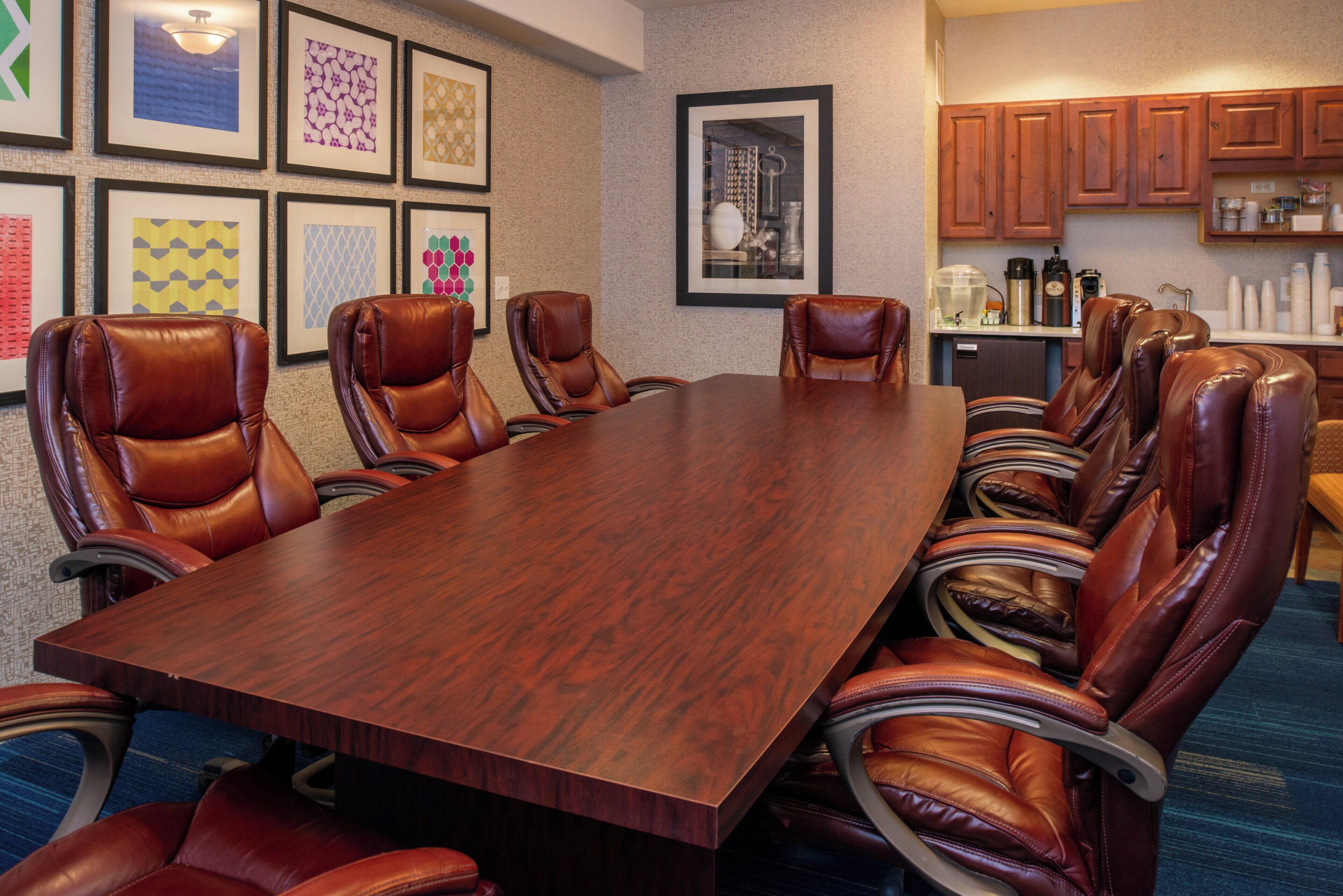 A conference room with a long wooden table and leather chairs, framed artwork on the walls, and a kitchenette area.