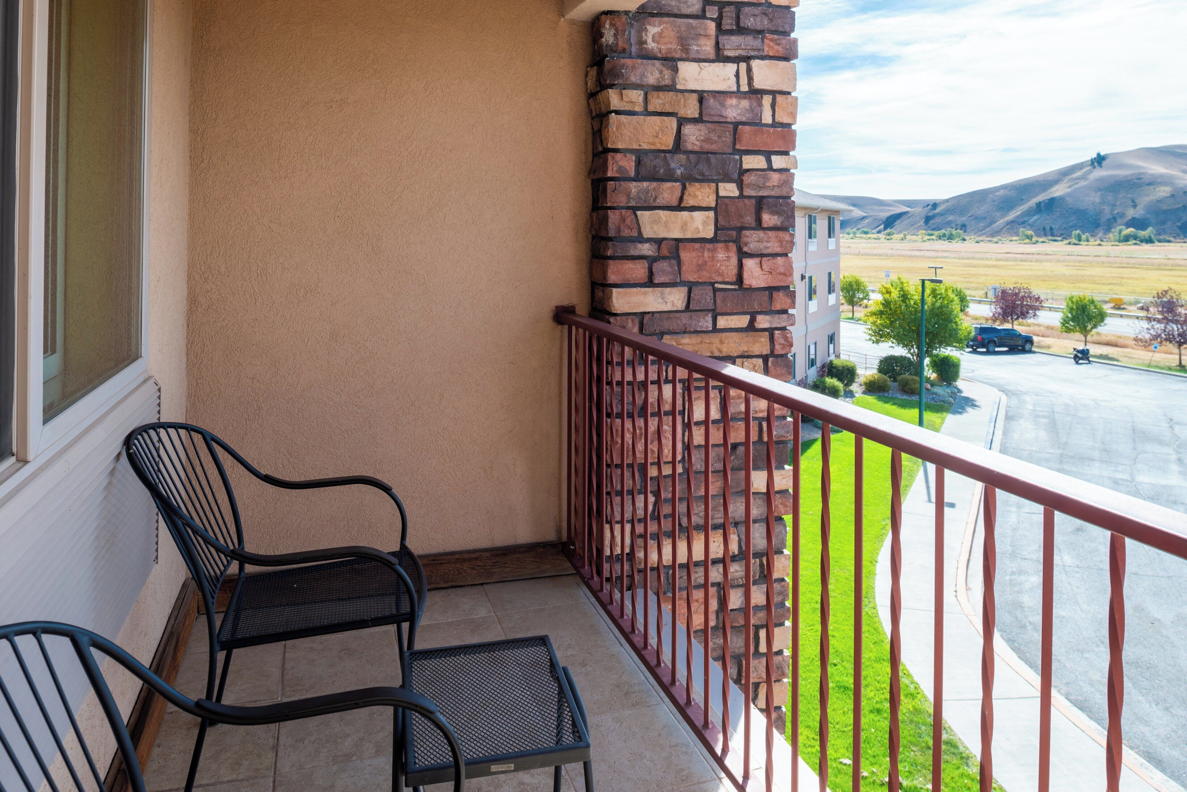 A balcony with two metal chairs, a stone wall, and a view of a road and distant hills.