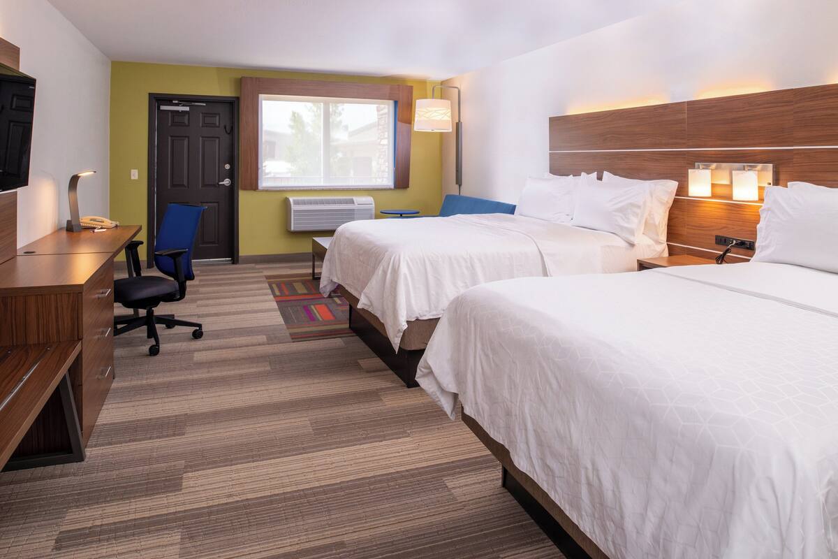 A hotel room with two beds, a desk with a chair, a television, and a window with a view of trees.