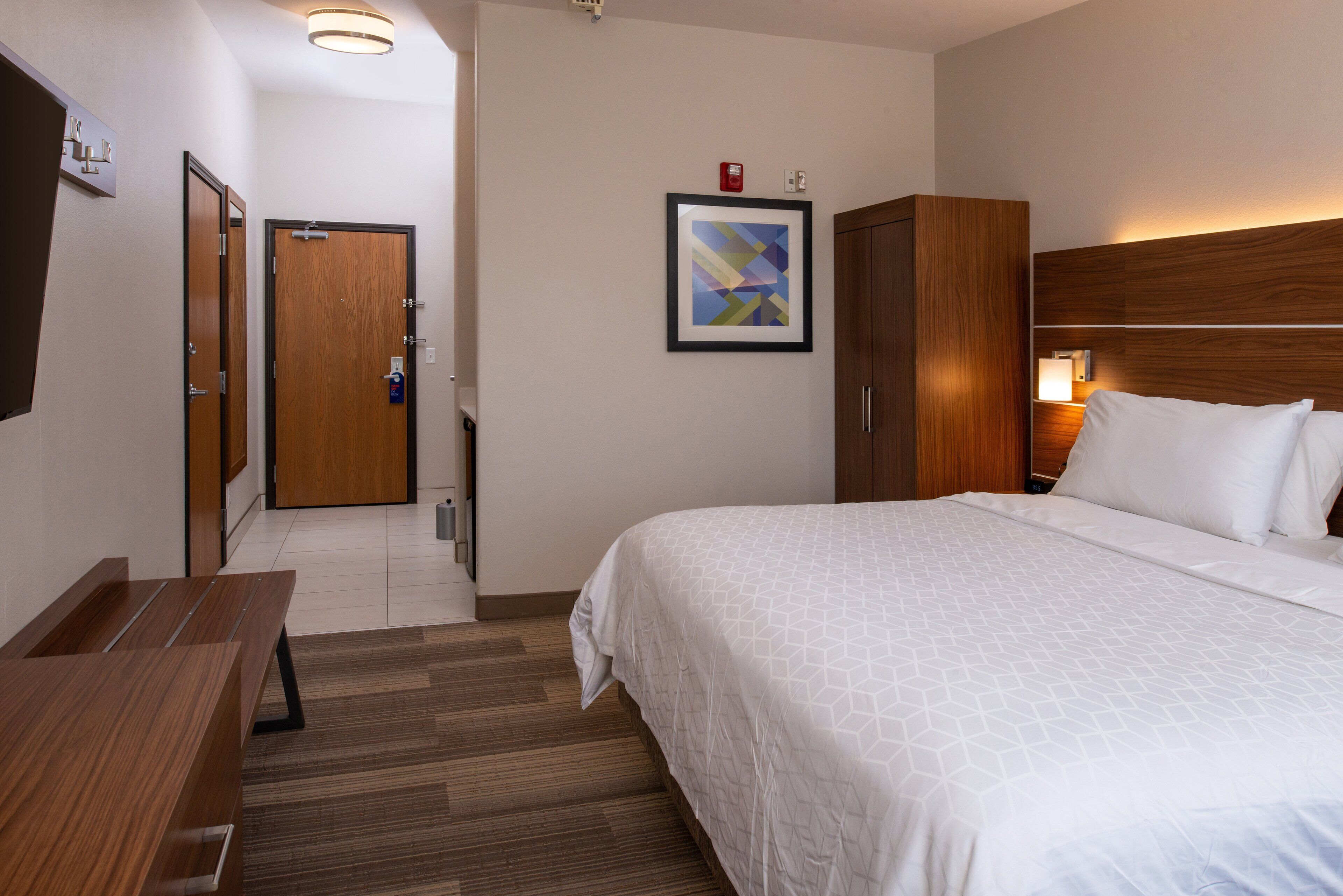 Una habitación de hotel con una cama, un escritorio de madera, un cuadro en la pared y una puerta que da a otra habitación.