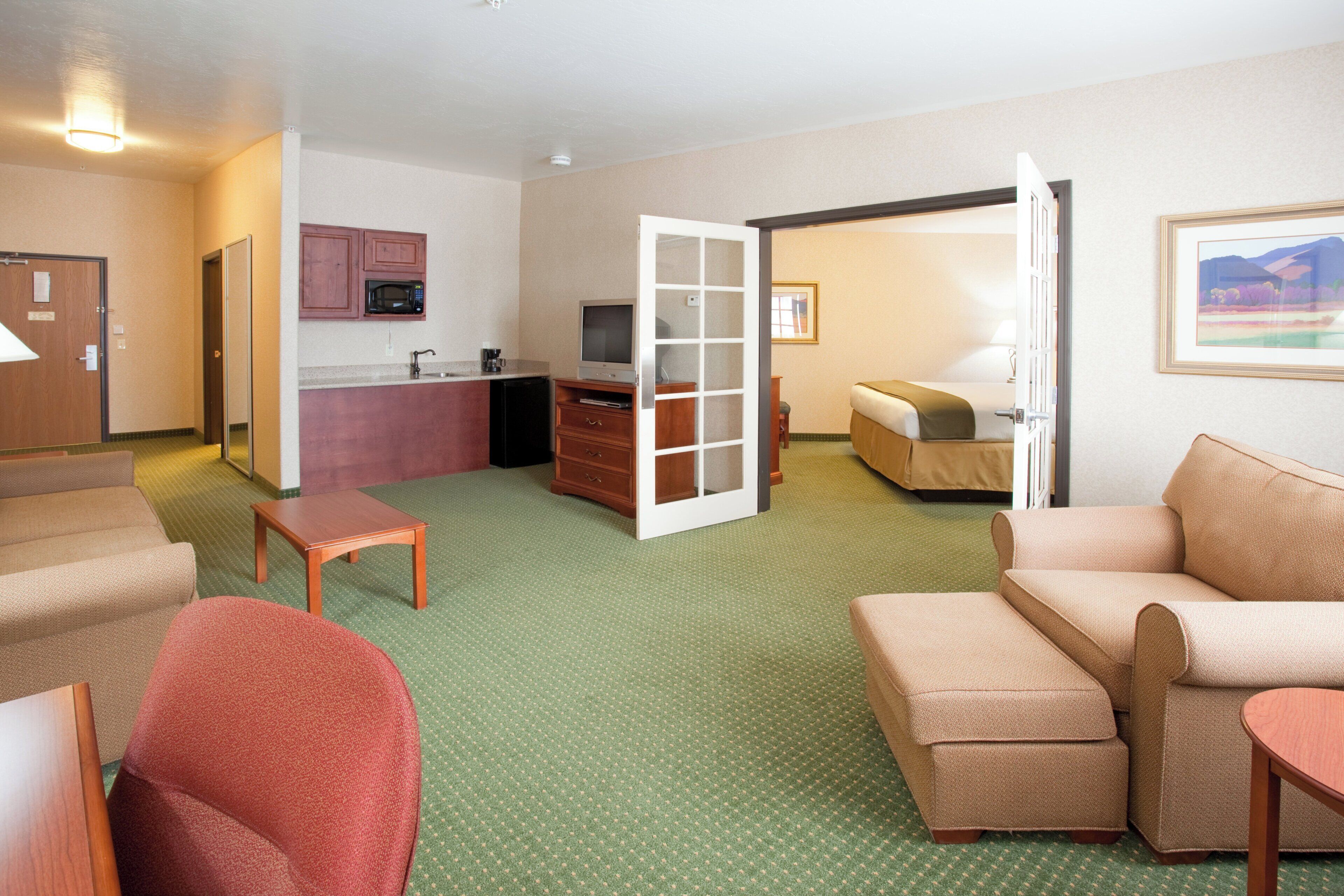 A hotel room with a kitchenette, a television, a bed, a seating area with a sofa and armchairs, and a carpeted floor.