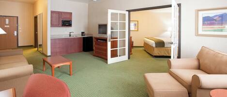A hotel room with a kitchenette, a television, a bed, a seating area with a sofa and armchairs, and a carpeted floor.