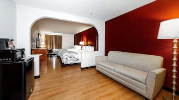 A hotel room with a wooden floor, a sofa, a desk with a lamp, a bed with a patterned bedspread, and a television.