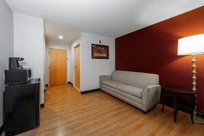 A hotel room with a wooden floor, a beige sofa, a small table with a lamp, a microwave, and a refrigerator.