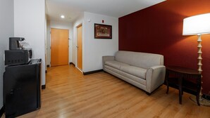 Quarto de hotel com piso de madeira, um sofá, uma escrivaninha com luminária e uma televisão.