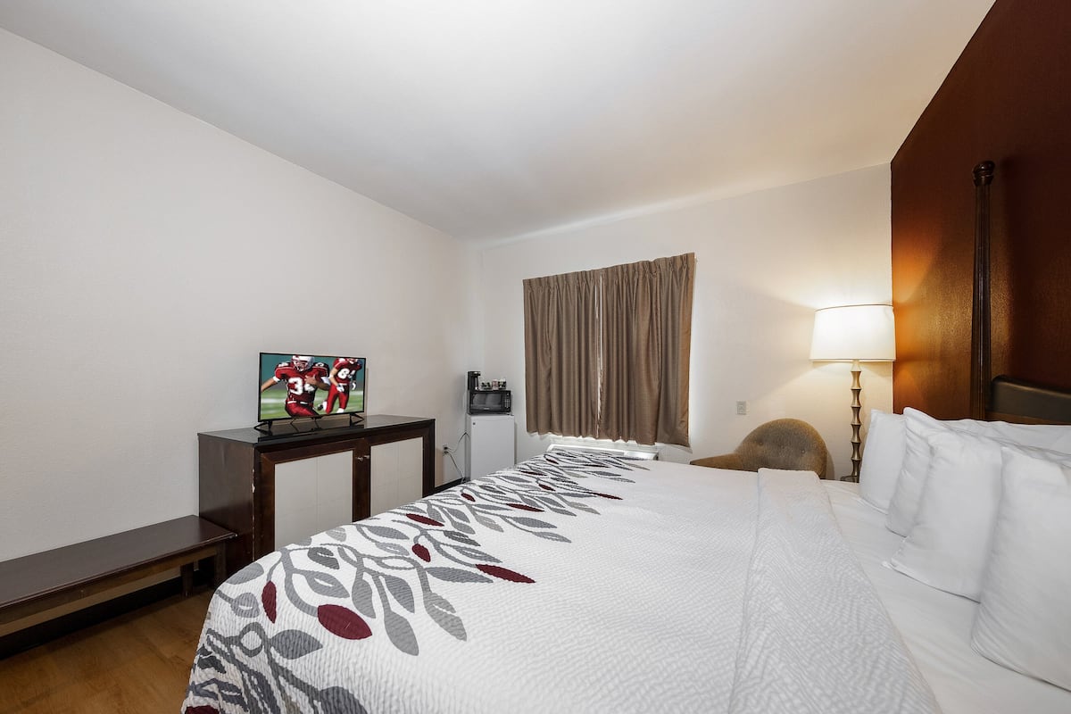 Ein Hotelzimmer mit Bett, Schreibtisch mit Fernseher, einem kleinen Tisch, einer Lampe und einem Fenster mit Vorhängen.