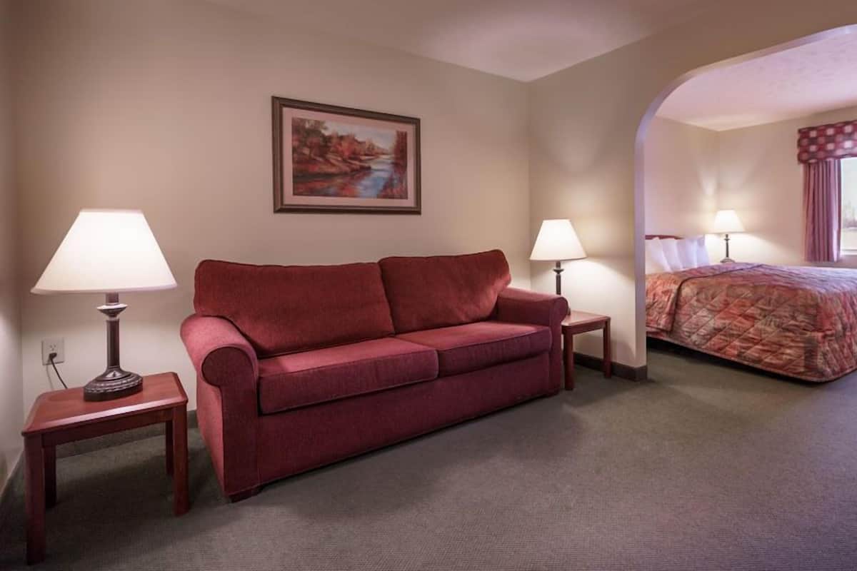 A hotel room with a red sofa, two bedside tables with lamps, a bed with a patterned bedspread, a painting on the wall, and a window with curtains.
