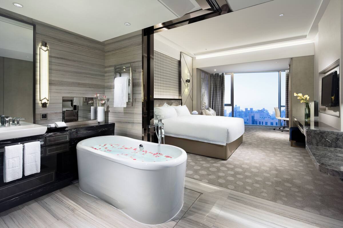Ein modernes Hotelzimmer mit einer großzügigen Badewanne, einem Bett, einem Badezimmer mit Waschbecken und Spiegel sowie Blick auf die Stadt.