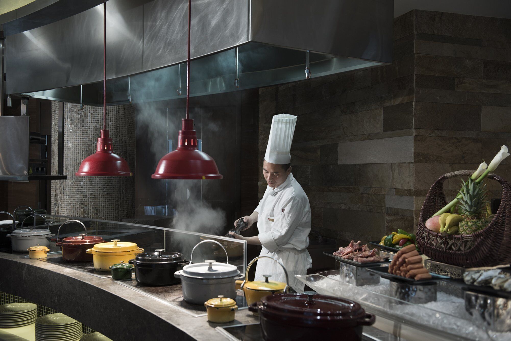 A chef in a professional kitchen with stainless steel appliances, hanging red pendant lights, and a variety of cooking pots and fresh produce on the counter.