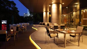 Un espace extérieur aménagé pour les repas, avec des tables et des chaises, une terrasse en bois et un bâtiment en verre en arrière-plan.