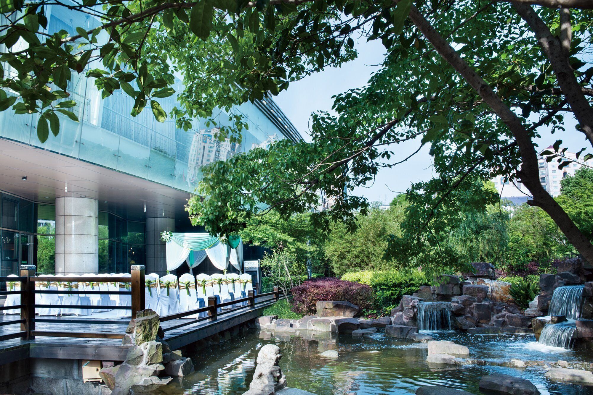 Un bâtiment moderne à la façade vitrée, un pont agrémenté de chaises blanches drapées et une petite cascade dans un jardin paysager.