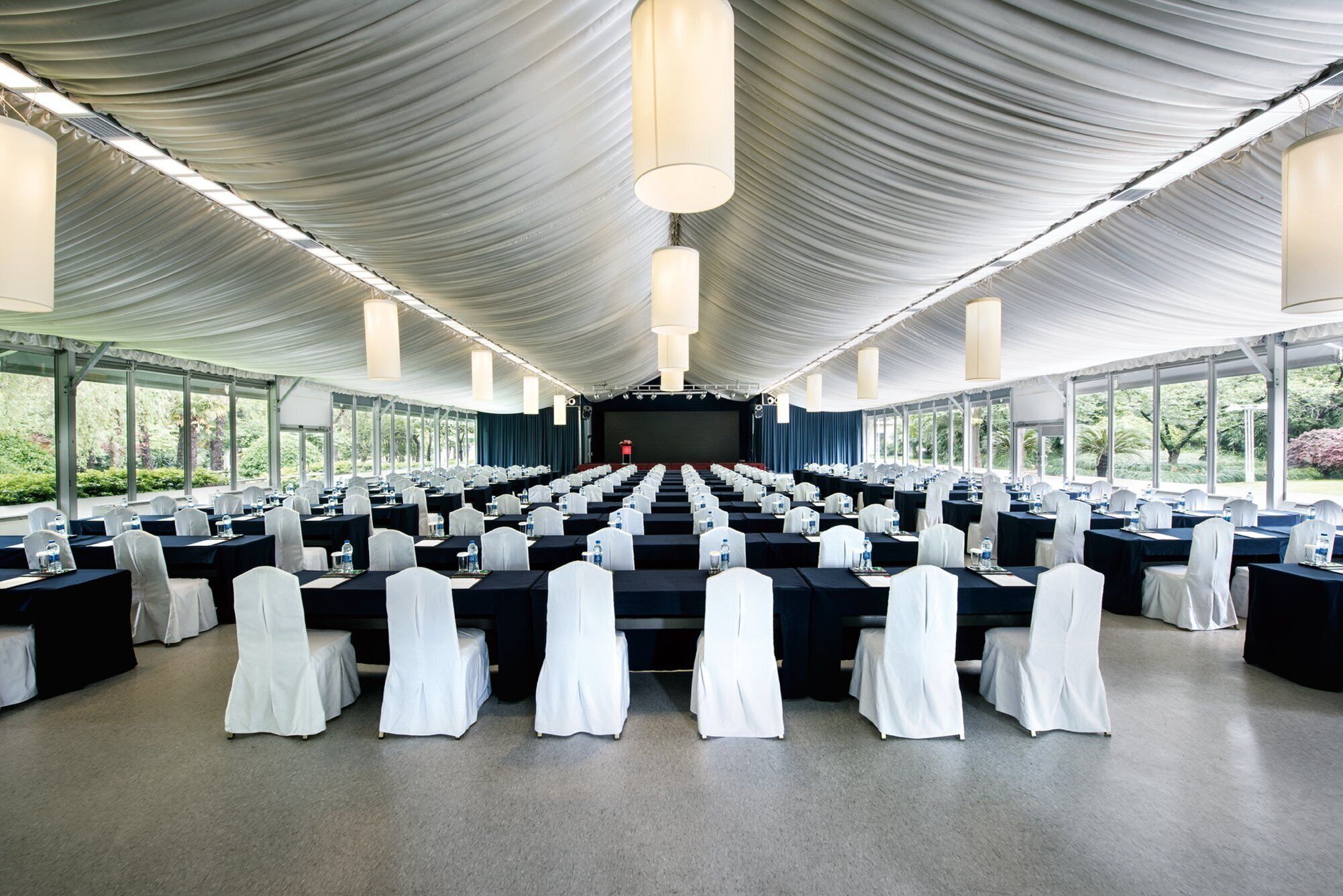 A large conference room with rows of tables and chairs, a stage at the front, and large windows along the sides.