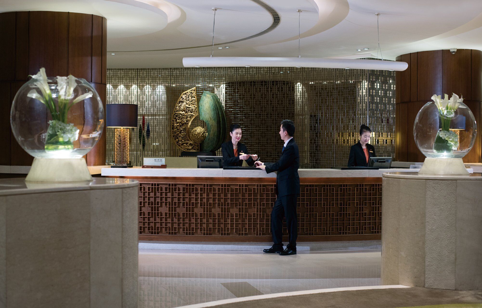 A hotel reception area with a modern design, featuring a reception desk, two receptionists, and decorative vases with flowers.